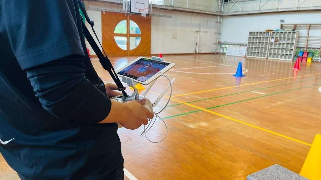 体育館内でドローン送信機を操作し、タブレットで飛行画面を確認している受講の様子